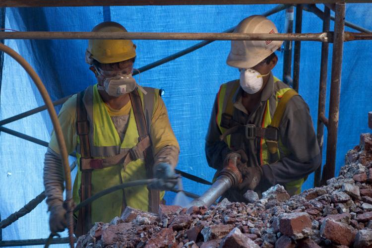 Two construction workers in hard hats and safety vests demolish a brick wall with a jackhammer inside Pagoda Palace.