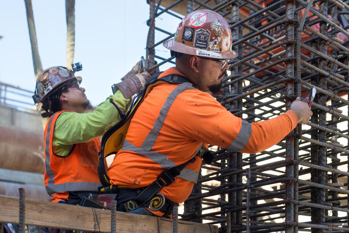 Two construction workers in safety gear secure rebar cage components on a building mezzanine level.
