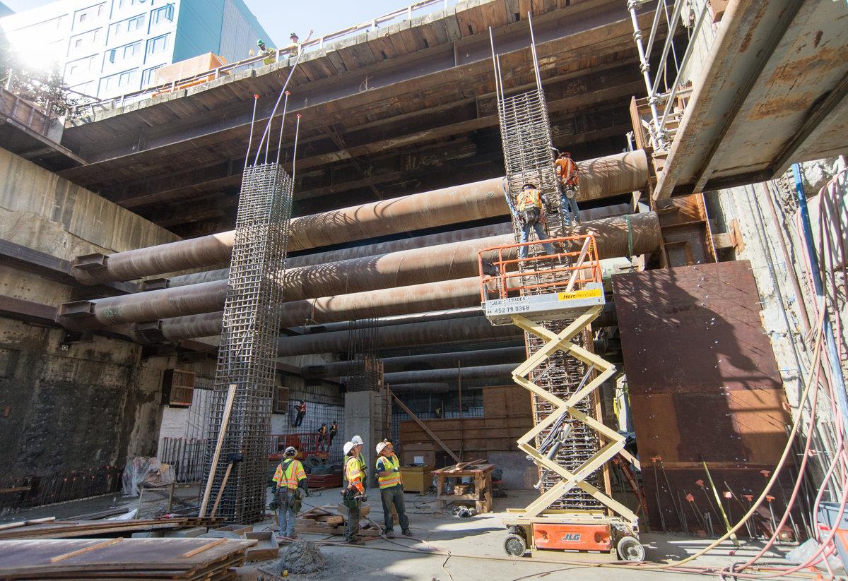 Construction workers on scissor lift installing large steel pipes under elevated highway structure for Central Subway project.