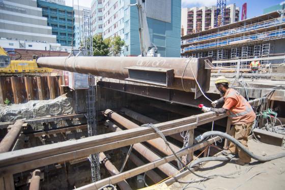 Worker guides large steel waler being lifted from underground construction pit in urban area.