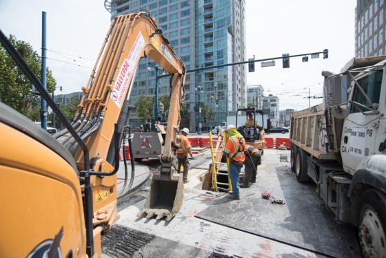 Construction excavator and workers at urban street intersection with transit vehicles and high-rise buildings.