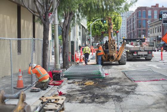 Construction crew works on street excavation with heavy equipment, safety barriers, and traffic cones at urban intersection.