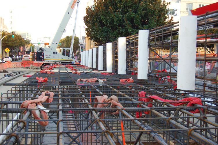 Construction site with rebar cage, white support columns, crane, and safety fencing on urban street.