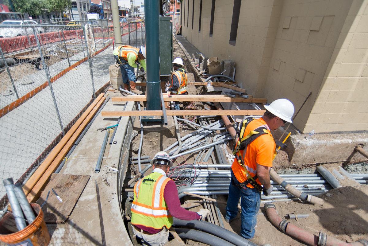 Construction workers install underground transit infrastructure including conduits and drainage systems at an urban construction site.