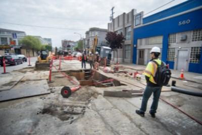Worker in safety gear observing utility vault installation on 4th Street construction site.