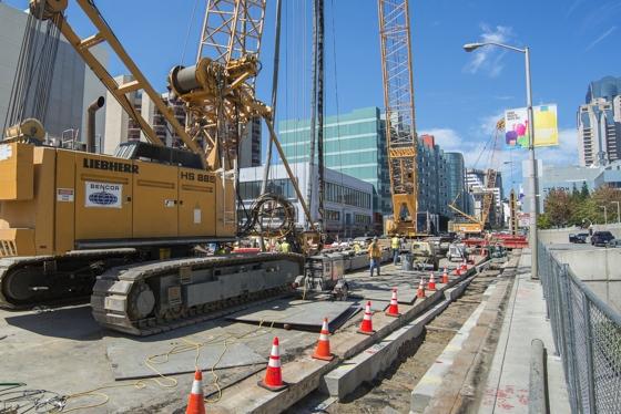 Yellow Liebherr hydromill excavator on 4th Street construction site with tall cranes and urban buildings in background.