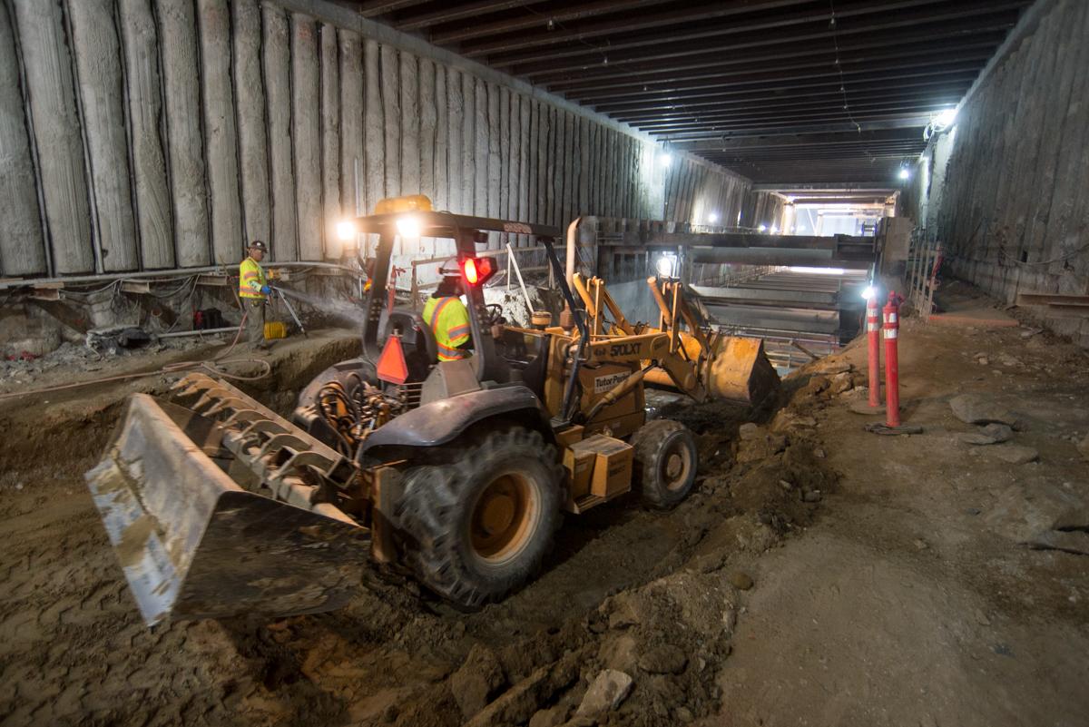 Workers operate heavy equipment to excavate and grade soil inside an underground transit station construction site.