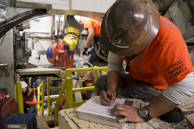 Worker in orange Central Subway shirt documents tunnel boring machine maintenance underground.