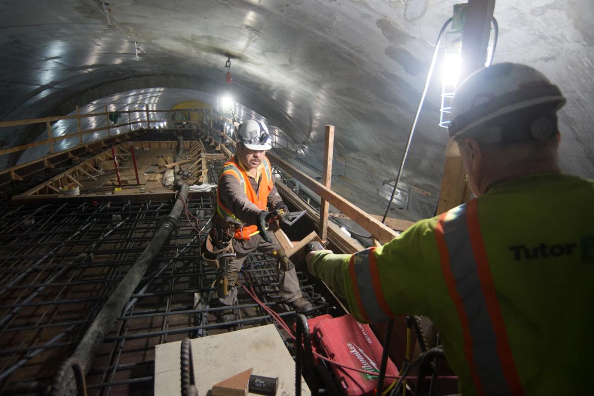 Workers transfer concrete formwork inside underground transit tunnel with steel reinforcement visible.