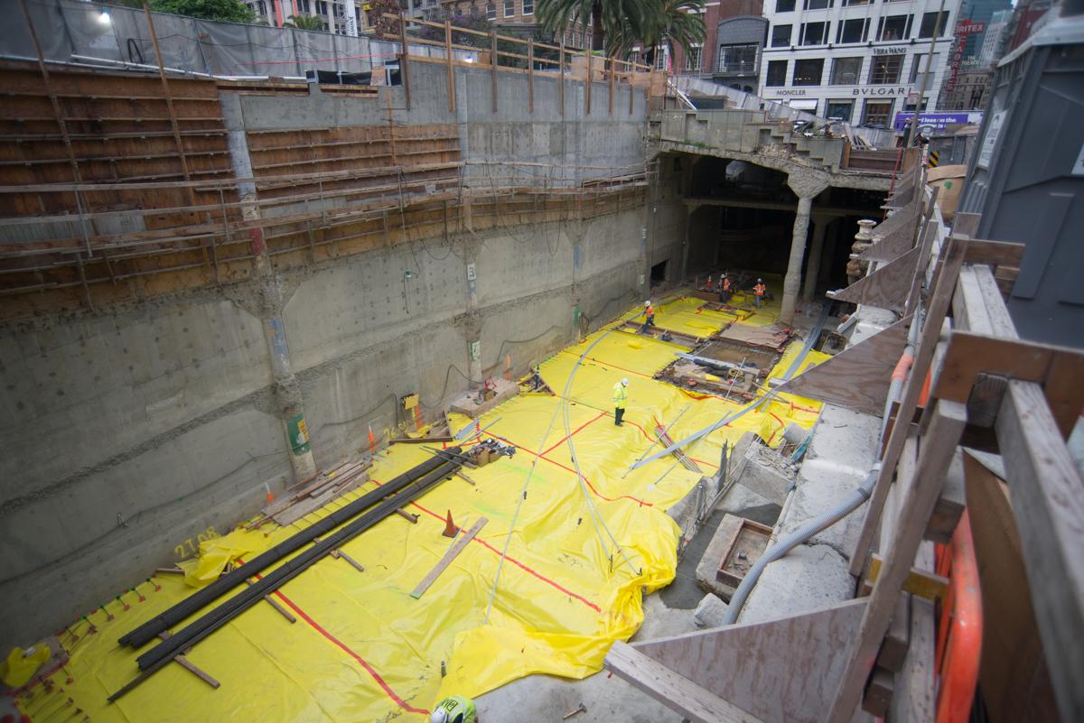 Underground construction site with yellow waterproofing material and rebar cages at station entrance excavation.