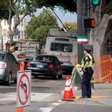 Construction work site with traffic control barriers, worker in safety vest, and street closure signage on urban street.