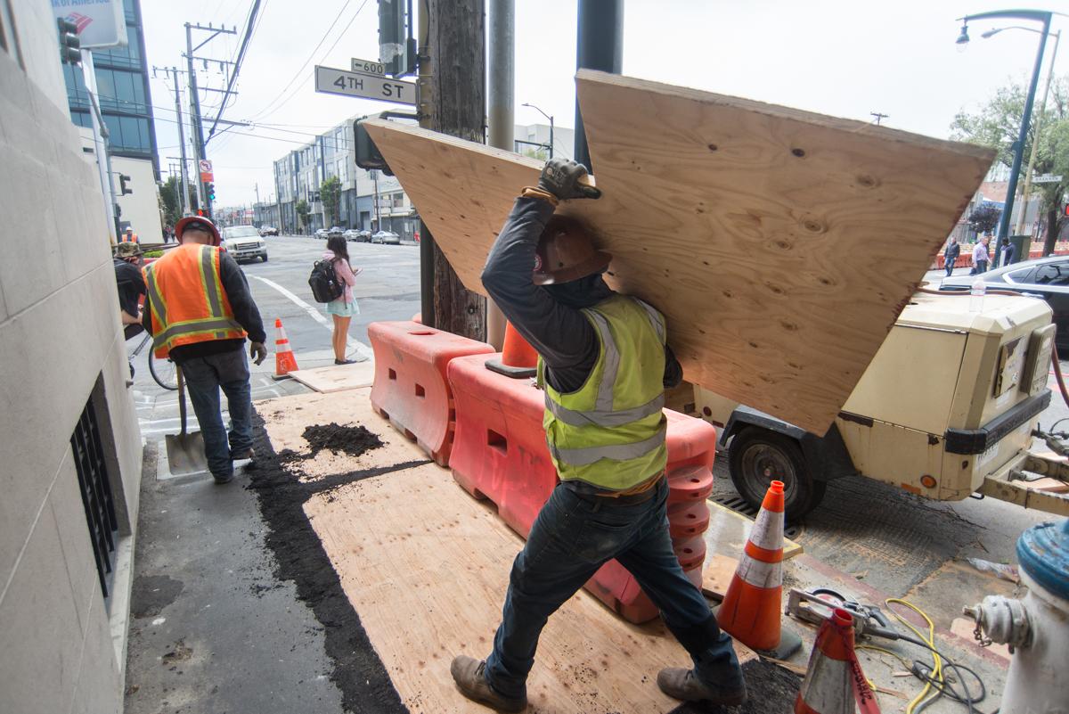 Worker in safety vest hoisting plywood to cover utility trenches on sidewalk at 4th Street construction site.