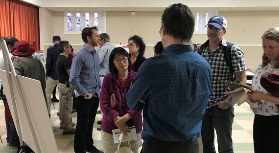 Group of people gathered indoors at an open house meeting, standing near informational boards and discussing transit project details.