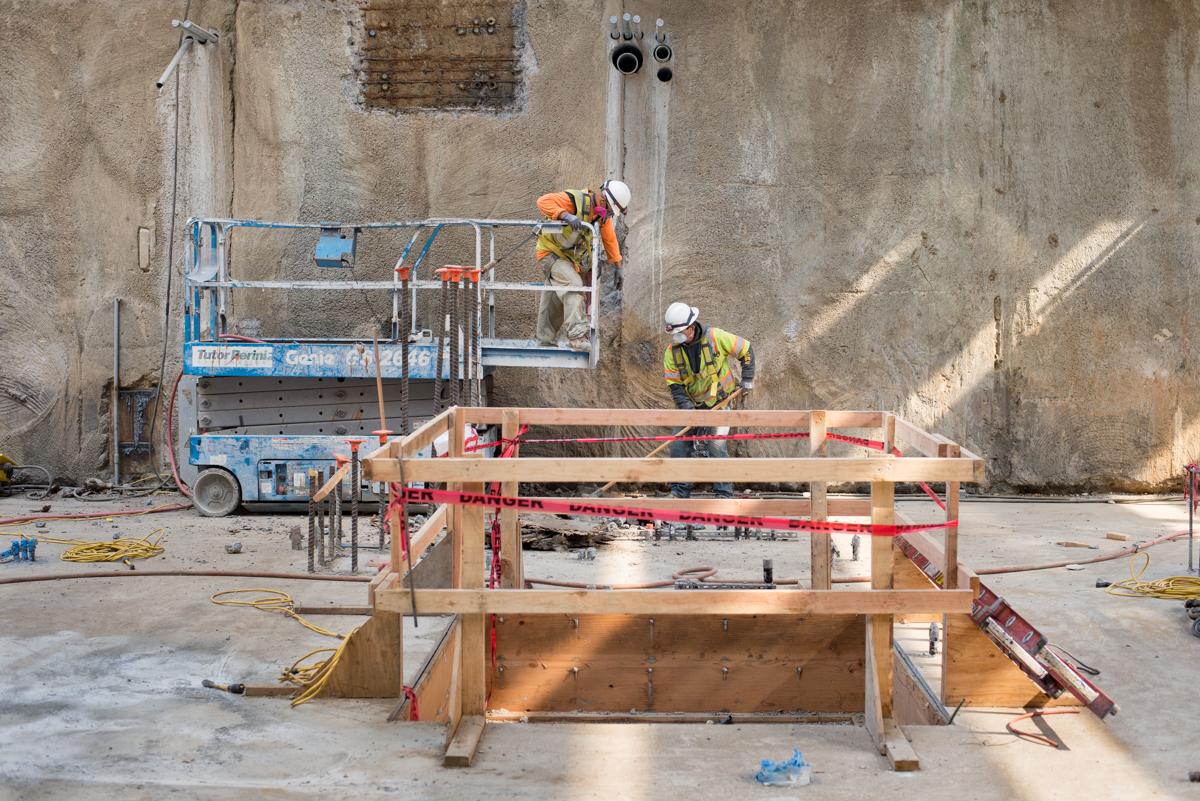 Construction workers on concrete platform with equipment and rebar at transit station headhouse site.