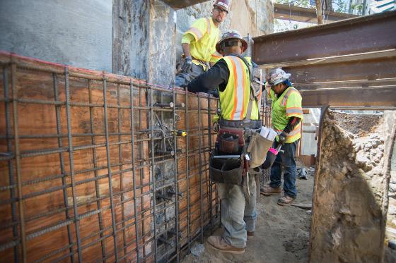 Construction workers in safety vests installing rebar for structural walls at an urban intersection.