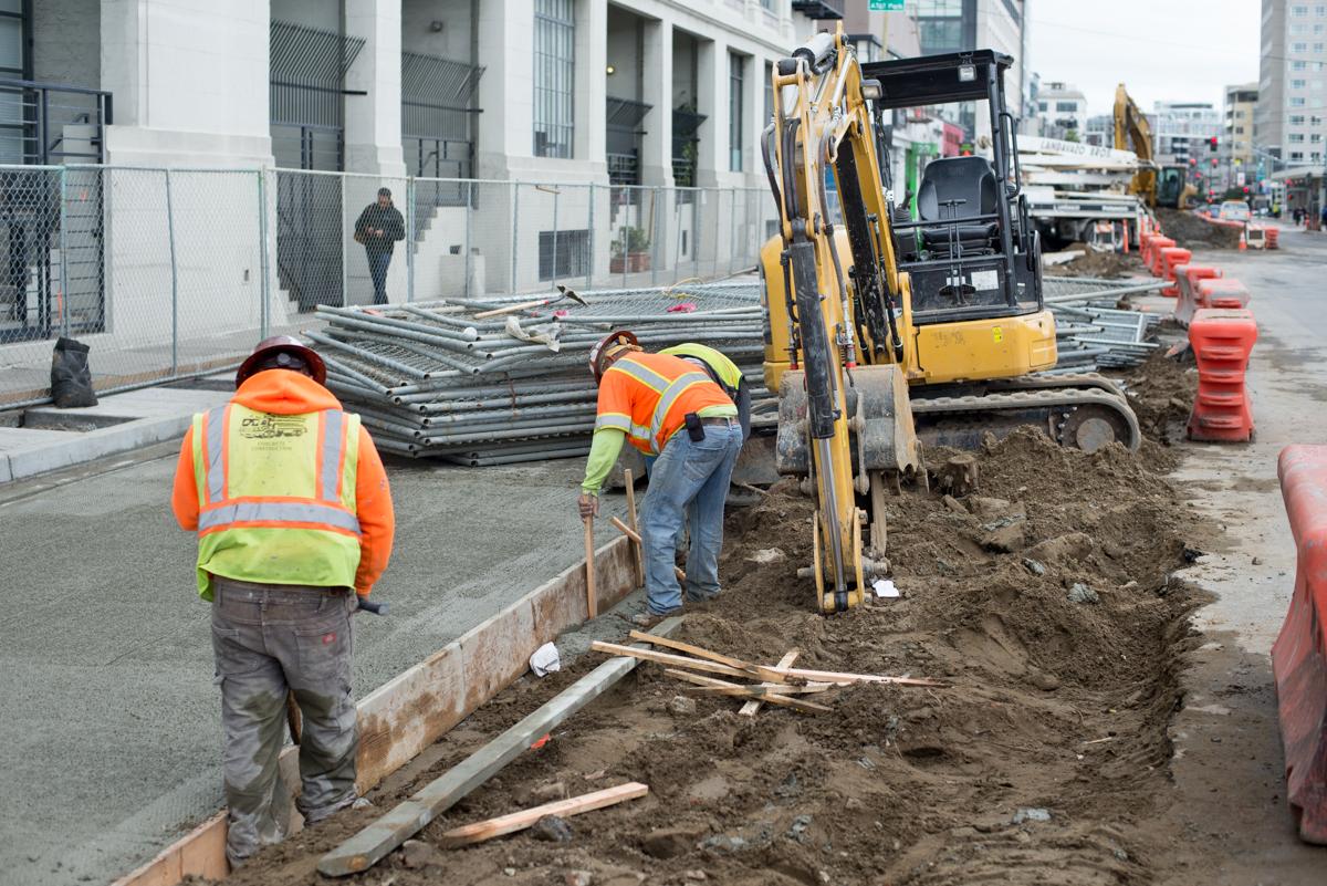 Construction workers in safety vests operate excavation equipment on 4th Street during utility work.