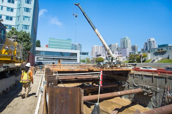 Construction site with crane positioned over excavation pit, worker in safety gear, city skyline in background.