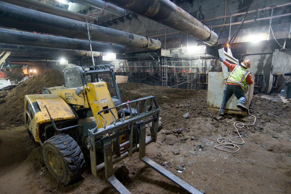 Worker in safety vest receiving steel lift box at underground station excavation site with heavy equipment.