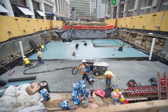 Construction workers installing waterproofing on underground transit station roof deck surrounded by yellow barriers.