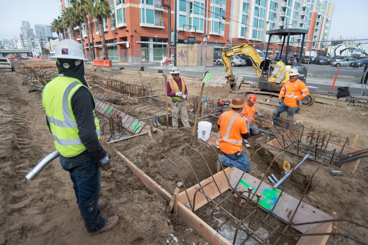 Construction workers in safety vests install utility conduits in trenches at a transit project site near urban buildings.