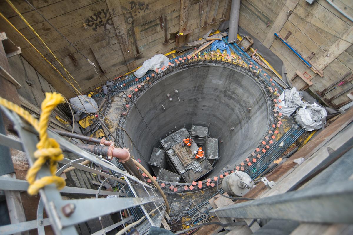Overhead view of deep circular shaft with workers, equipment, and yellow safety rigging visible inside construction site.