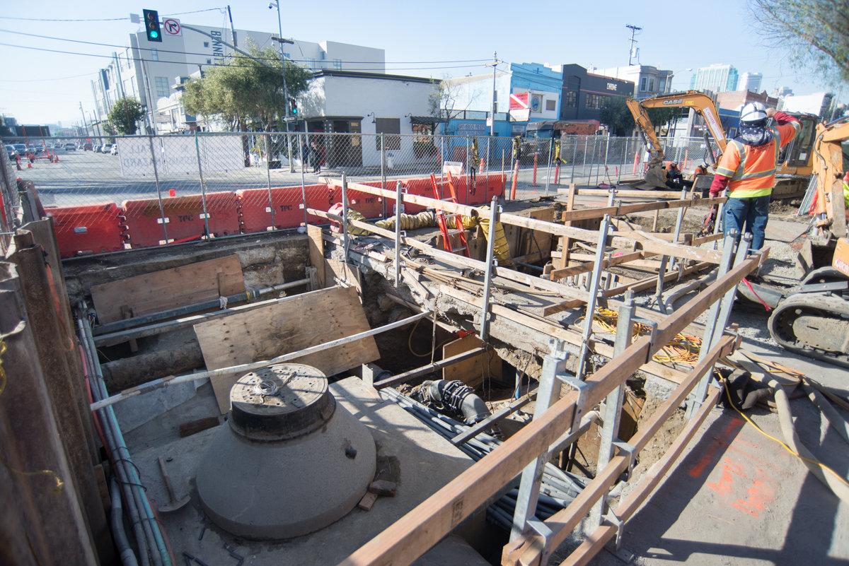 Construction site with underground utility work, excavation pit, and sewer infrastructure on urban street.