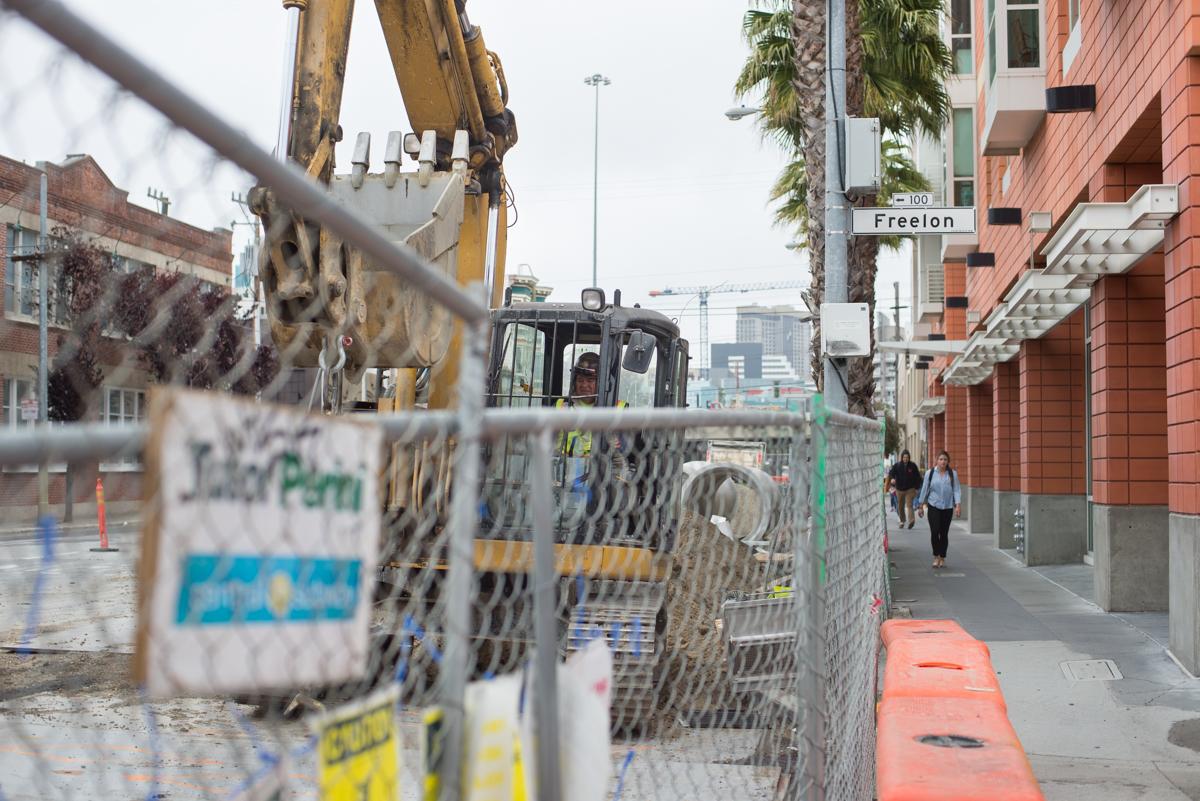Construction site at Freelon Alley with excavator, fenced work area, and workers on adjacent sidewalk.