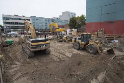Construction site with yellow excavators and wheel loader at Yerba Buena Moscone Station, urban buildings in background.