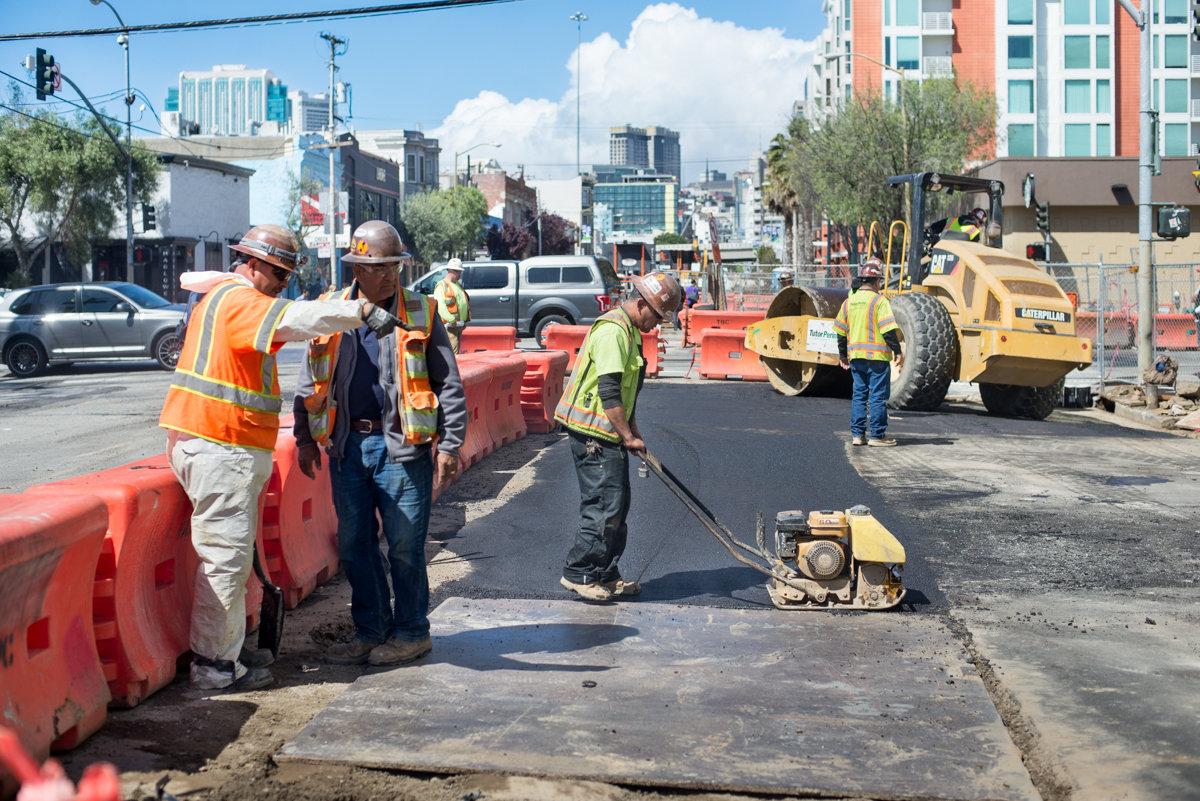 Construction workers lay asphalt on 4th Street with heavy equipment and traffic barriers visible.