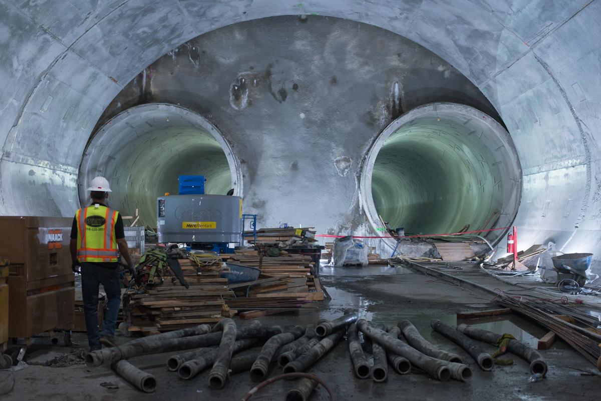 Worker in safety gear inside tunnel with large pipes and construction materials during transit project.
