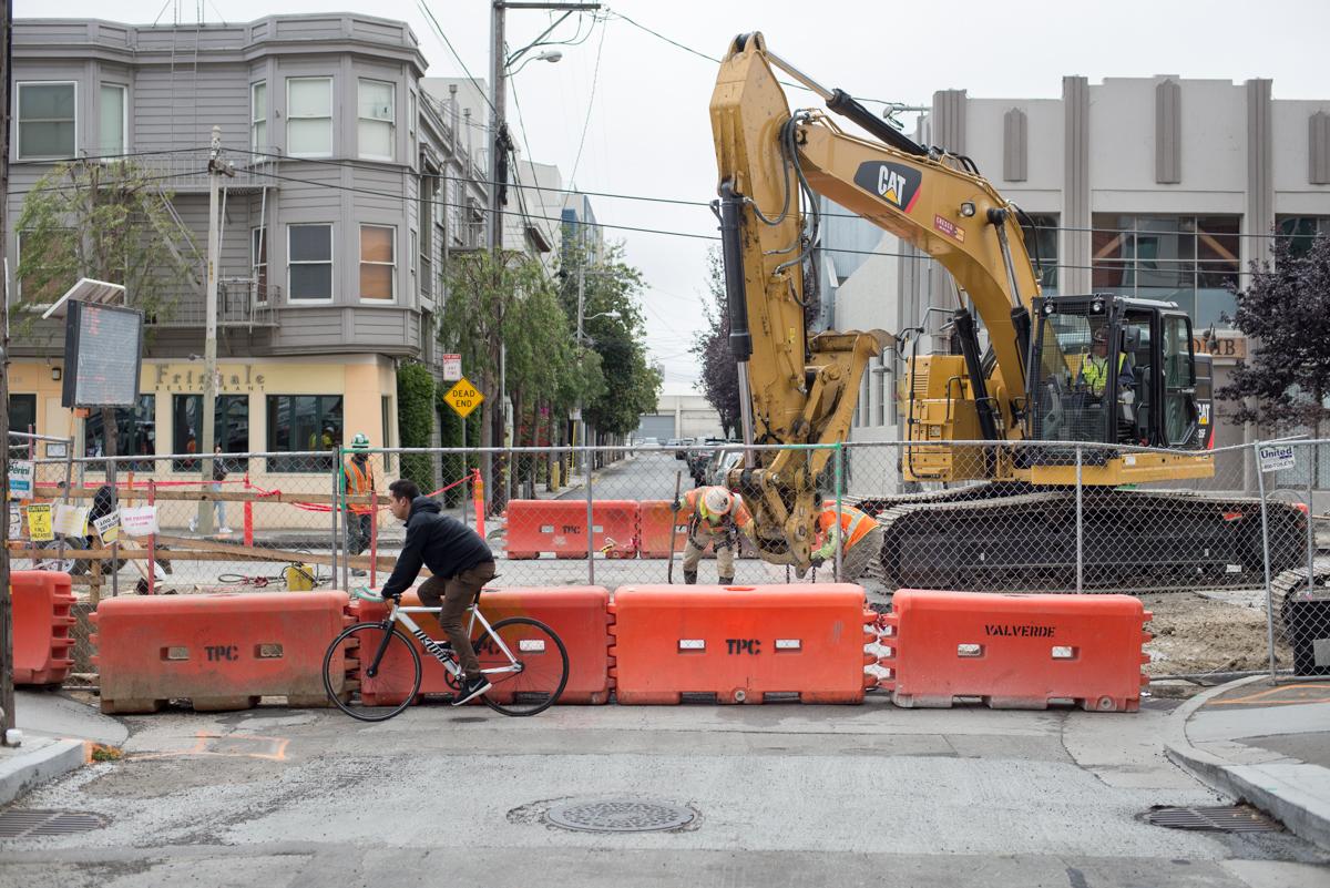 Cyclist navigating around orange barriers at construction site with excavator on Townsend Street.