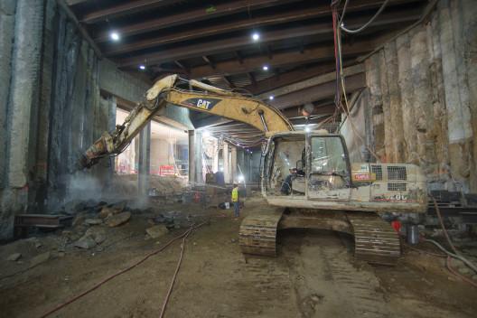 Excavator with jackhammer attachment removing concrete from underground transit station walls.