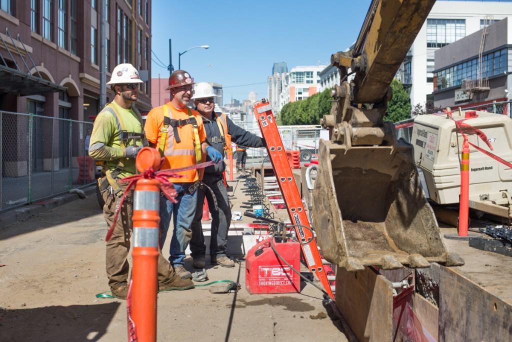 Three construction workers in hard hats and safety vests stand near excavation equipment and an orange traffic cone on an urban street.