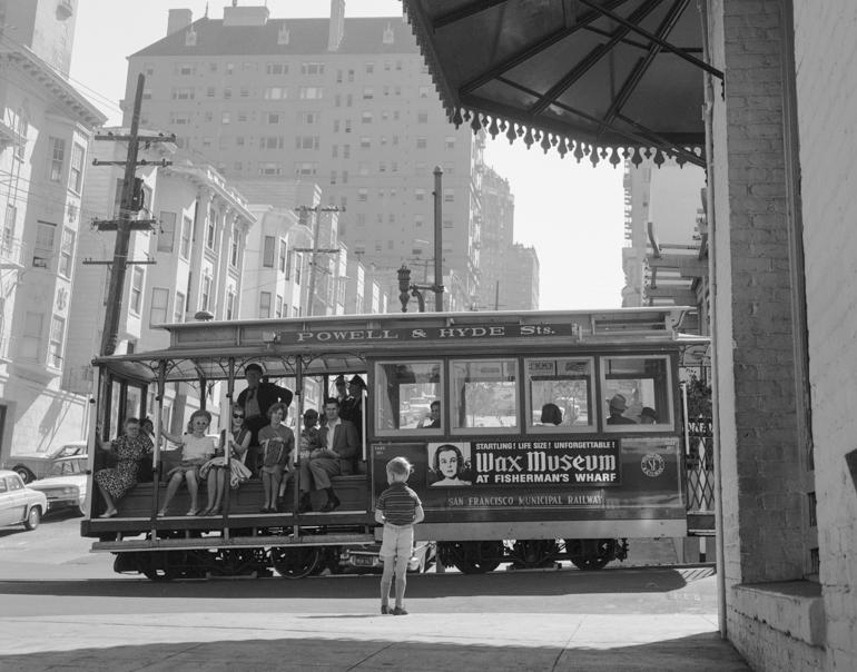 Child standing on street corner watching Powell and Hyde cable car pass Washington and Mason Streets, San Francisco, 1965.