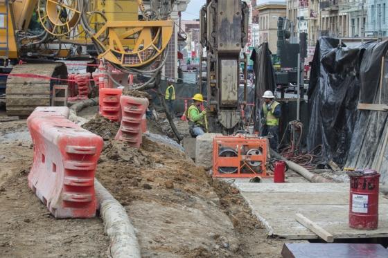 Construction site with yellow hydromill equipment, red barriers, and workers in safety vests on Stockton Street.