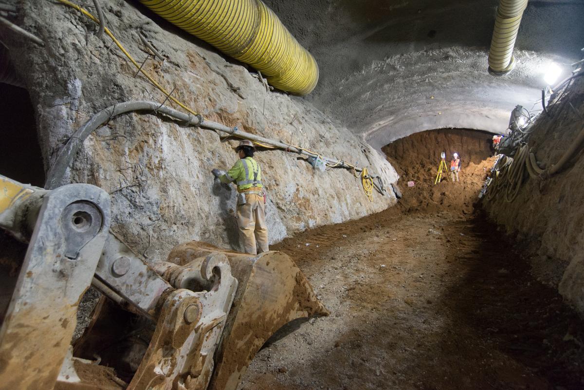 Worker in safety vest taking measurements inside underground transit tunnel excavation site with concrete walls and equipment visible.