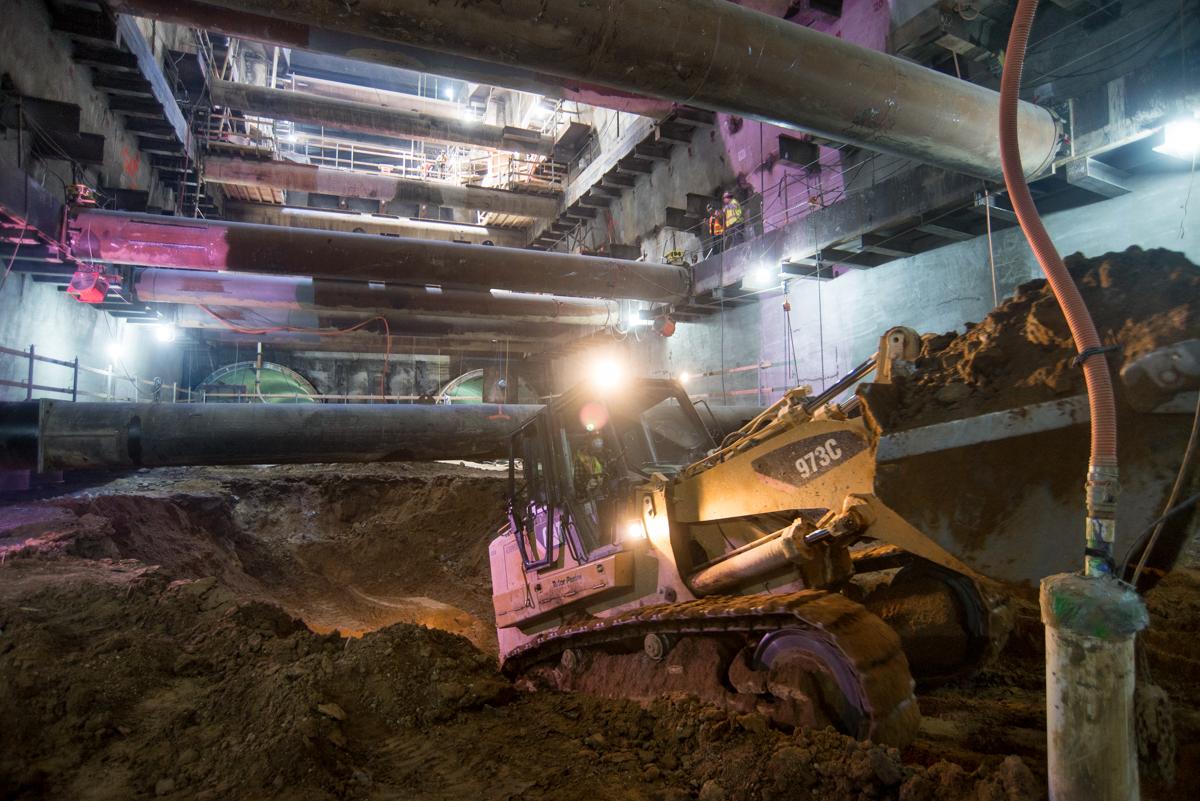 Bulldozer excavating soil from underground station construction site with industrial lighting and steel framework overhead.