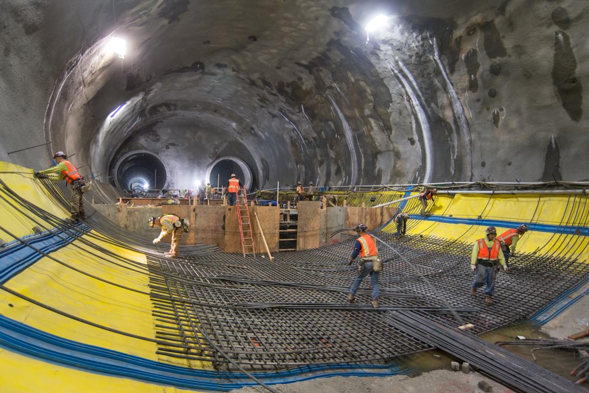 Workers in safety vests install reinforcing steel and waterproofing materials inside a tunnel under construction.