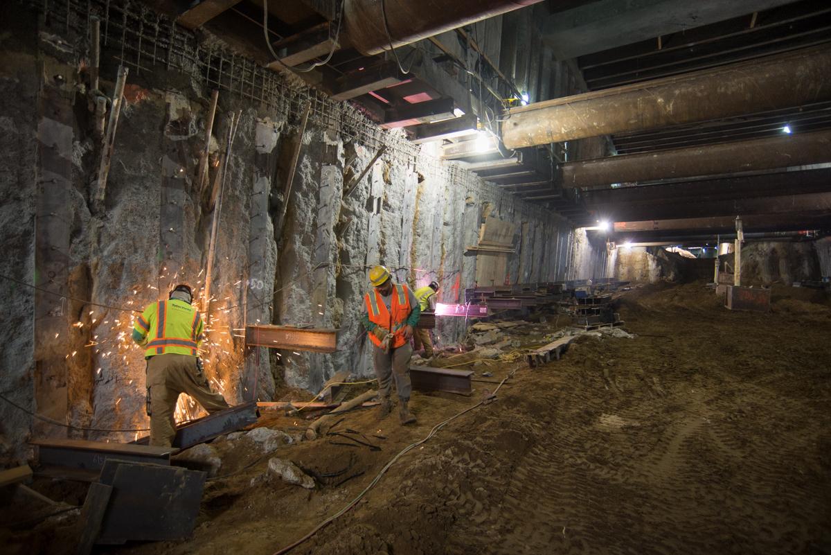 Workers in safety vests perform welding and steel reinforcement installation in an underground transit station tunnel.