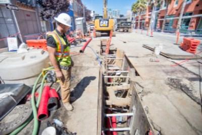 Worker in hard hat and safety vest at street construction site with excavator and utility work in progress.