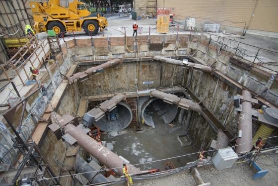 Excavation pit showing two large circular tunnel boring machine openings with water pooling at bottom, yellow loader visible above ground.