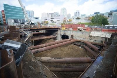 Temporary steel supports span excavation pit at Yerba Buena Moscone Station construction site with city skyline behind