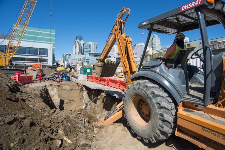 Construction worker operating backhoe to remove utilities at Yerba Buena/Moscone Station site with city skyline in background.