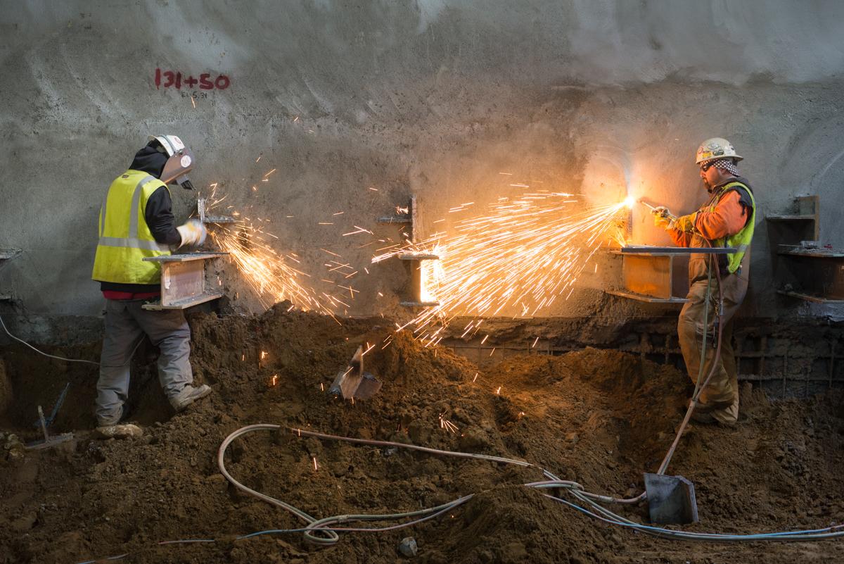 Two workers in safety gear use cutting torches on metal brackets in an underground construction site.