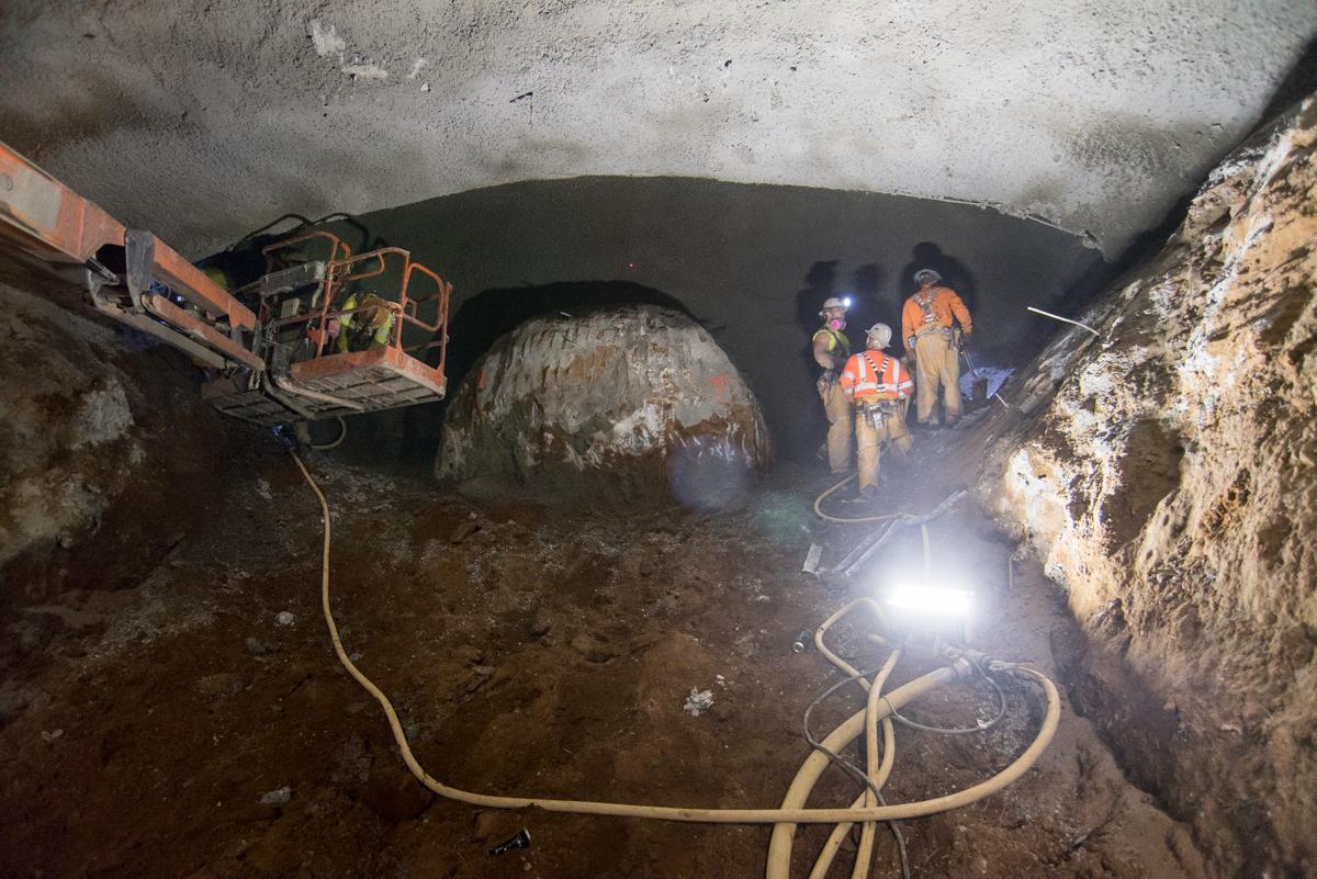 Workers in hard hats inside underground tunnel with concrete ceiling, equipment, and lighting cables during construction excavation.