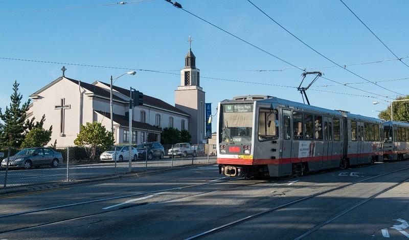 Muni M Ocean View light rail train stopped at 19th Avenue near a church with a steeple.