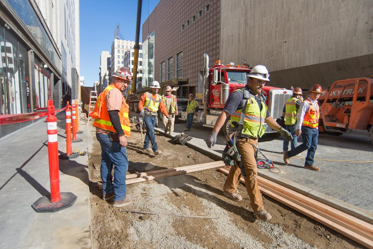 Construction workers in safety gear work on street excavation along an urban roadway with buildings and vehicles nearby.