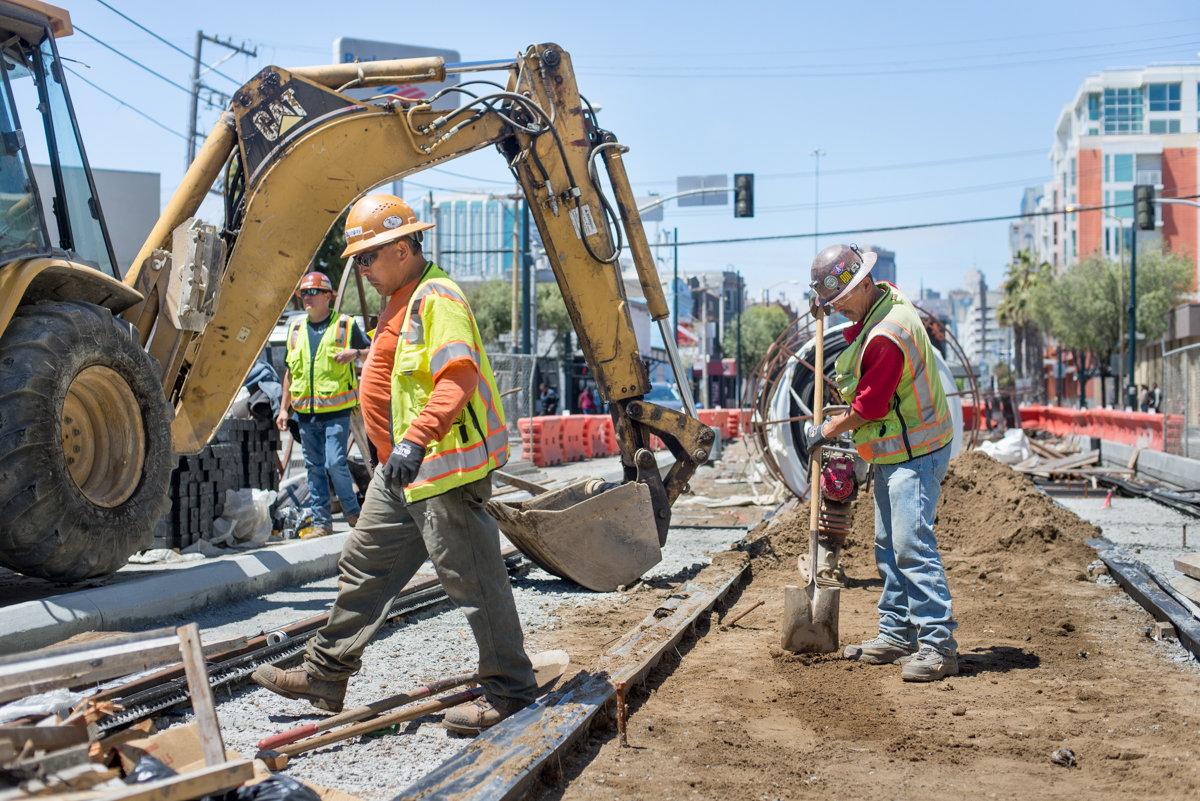 Construction workers operate an excavator to dig and backfill soil on a city street during transit infrastructure work.