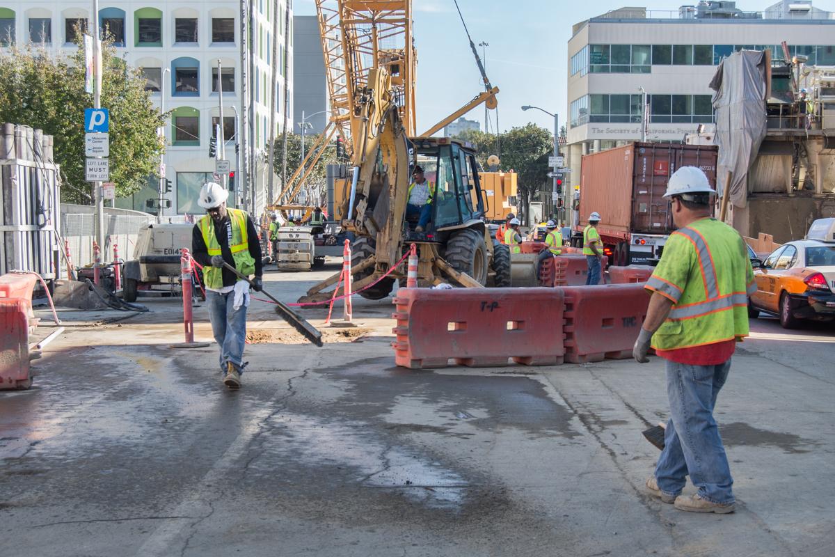 Construction workers in safety vests and hard hats work on a street with excavation equipment and barriers during underground station construction.