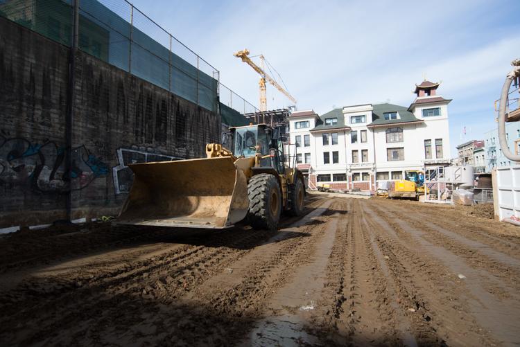 Construction equipment grading dirt at Chinatown Station site with buildings and crane in background.